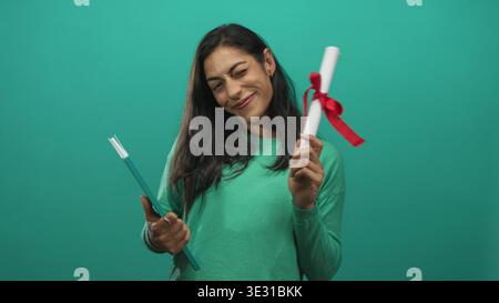 Woman smiling while holding a diploma and teal book in studio with hands visible and tilted head; graduation pride accomplishment. Stock Photo