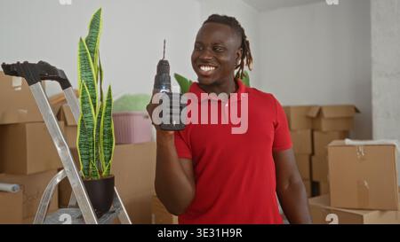 Man holding drill beside ladder and potted plant in building cluttered with moving boxes and stacked cartons, smiling broadly; home improvement joy. Stock Photo