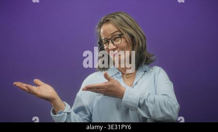 Woman gestures presenting open palm in studio beside purple seamless backdrop, wearing glasses and light blue shirt; warmth invitation. Stock Photo
