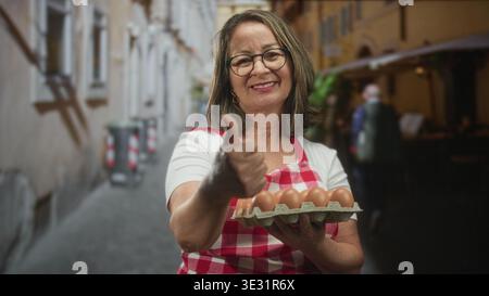 Hispanic woman with grey hair wears red checked apron, holds carton of eggs and shows thumbs up in street; home cooking pride. Stock Photo