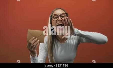 Young woman holding kraft envelope with outstretched hand and smiling gesture in studio; joy surprise greeting. Stock Photo