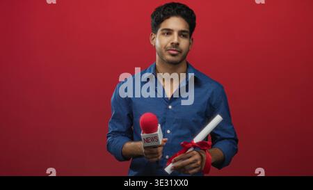 Man holding red news microphone and ribboned diploma with hands visible against a red backdrop in studio; thoughtful contemplation. Stock Photo