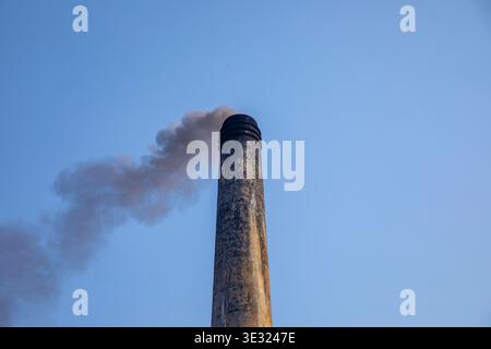 Thick black smoke rises from a brickfield chimney into the clear blue sky, illustrating the environmental impact of traditional brick production and a Stock Photo