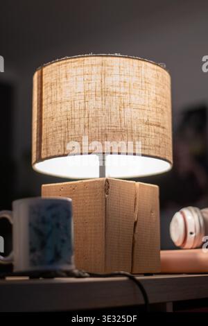 A linen lampshade emits a soft warm glow, lighting a wooden table with personal items in a calm home setting. The image captures a relaxing indoor Stock Photo