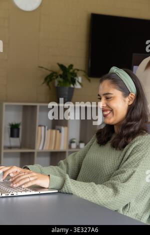 Asian woman typing on laptop keyboard at home wearing green knit sweater and headband, copy space Stock Photo
