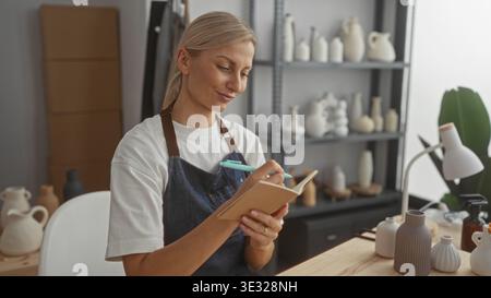 Woman potter writes in a notebook holding a pen near her lips while wearing an apron among ceramic vases on shelves in a pottery studio; thoughtful pl Stock Photo