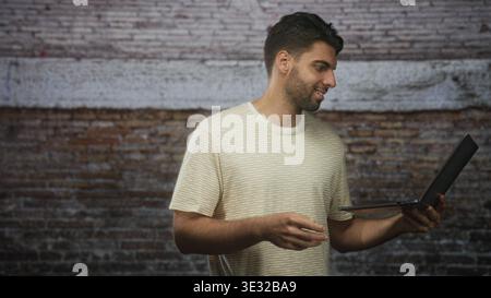 Hispanic man typing on laptop, fingers on keyboard and smiling, standing before a brick wall in studio; concentration productivity. Stock Photo
