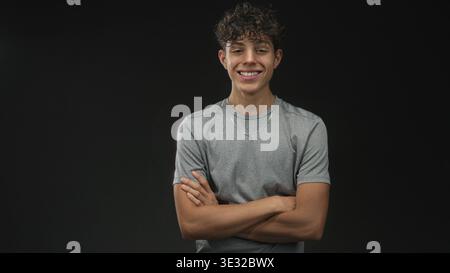 Boy wearing gray t shirt and silver necklace smiling with arms crossed in studio with black backdrop and soft lighting; youthful confidence. Stock Photo