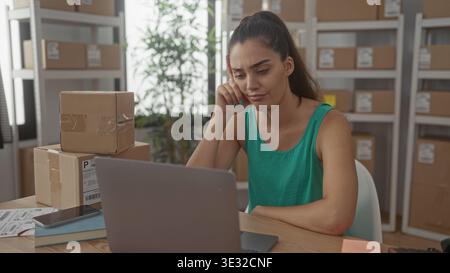 Woman rests hand on cheek scrolling on laptop beside stacked packages in building; ecommerce shipping anxiety. Stock Photo