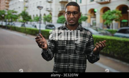 Young man holding car keys with hand to forehead on a city street near parked cars and hedges, wearing glasses and plaid shirt; frustration. Stock Photo