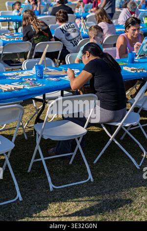 Lynn Haven, FL, USA. 20th Mar 2026. Community members gather outdoors at Sheffield Park for the free Art in the Park event, where the city provides canvases and paints for all ages to enjoy. In the foreground, a woman wearing a black t-shirt and grey sweatpants sits on a white folding chair at a blue-covered table, intently working on her small canvas. Numerous other attendees, including children and adults, sit along the rows of tables in the bright afternoon sunshine, channeling their creativity during the two-hour community gathering. Credit: Tosh Persons / Ģ����ý Live News Stock Photo