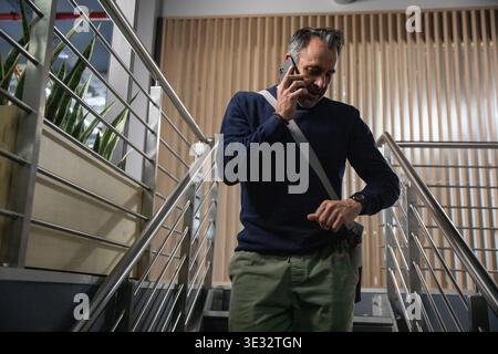 Middle-aged man descending stairwell, talking on smartphone and checking wristwatch near planter Stock Photo