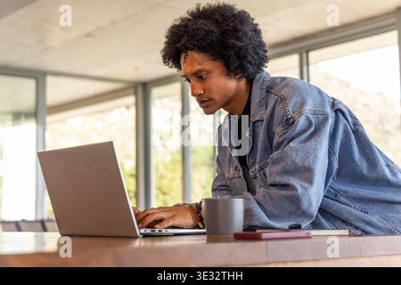 African-American male leaning, working on laptop at table with denim jacket wristbands phone mug Stock Photo