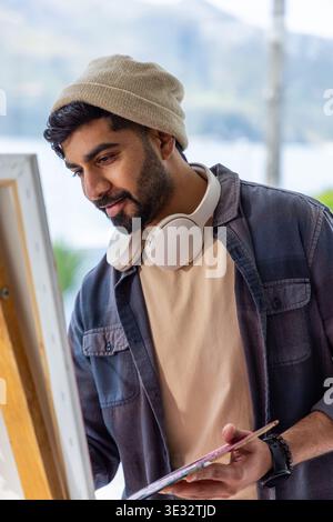 Indian man painting at easel in studio near window holding palette and wearing beanie Stock Photo