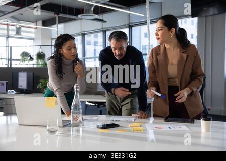 Diverse coworkers in office attire leaning and pointing at charts around table in open office Stock Photo