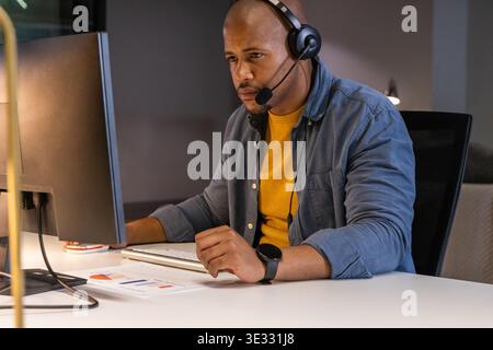 African American man working at desk in office, wearing headset and using monitor and keyboard Stock Photo