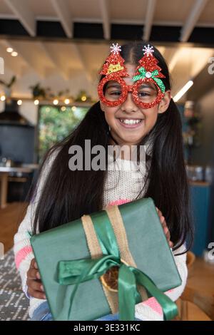 Asian child girl smiling and holding green-wrapped gift in kitchen wearing glittery red glasses Stock Photo