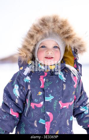 A warm‑clothed child stands in bright snow wearing a patterned winter jacket and fur‑lined hood Stock Photo