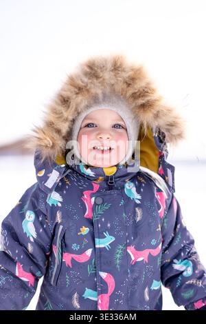 A warm‑clothed child stands in bright snow wearing a patterned winter jacket and fur‑lined hood Stock Photo
