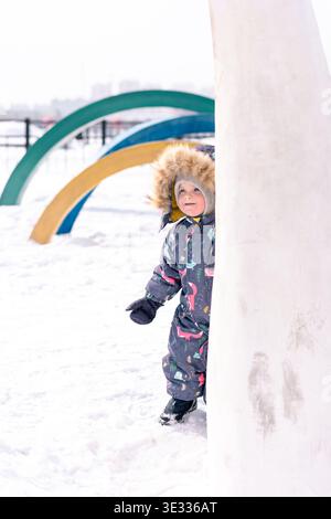 A child in a colorful winter snowsuit peeks playfully from behind a snowy structure Stock Photo