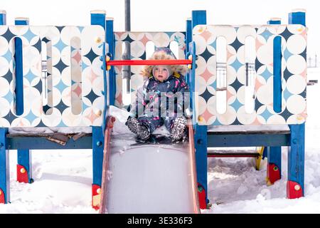 A child in a colorful winter snowsuit slides down a snowy playground slide outdoors Stock Photo