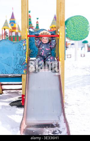 A child in a colorful winter snowsuit slides down a snowy playground slide outdoors Stock Photo