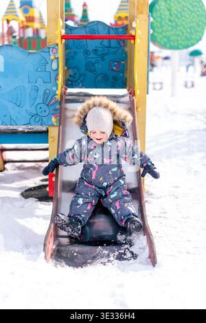 A child in a colorful winter snowsuit slides down a snowy playground slide outdoors Stock Photo