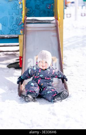 A child in a colorful winter snowsuit slides down a snowy playground slide outdoors Stock Photo