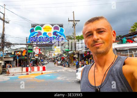 Handsome tourist male traveler man travels and takes selfie on tourist street road with people cars palm trees promenade shops bars and restaurants in Stock Photo