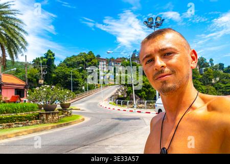 Handsome tourist male traveler man travels and takes selfie on tourist street road with people cars palm trees promenade shops bars and restaurants in Stock Photo