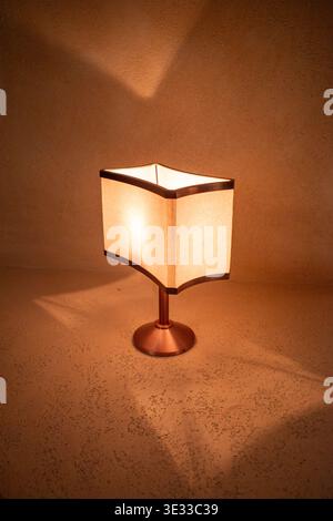 Warm light casts soft shadows from a square lamp. Minimalist design features cream-colored fabric shade. Metal base and stem offer sleek, modern contr Stock Photo