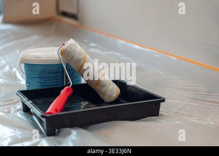 A paint roller and tray set next to a bucket of paint, prepared for a home renovation project. The floor is covered with protective plastic. Stock Photo
