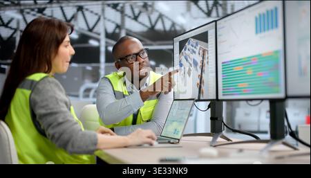 Young African Engineer Uses 3D Software On A Desktop To Optimize Factory Layout. Two Managers, A Female And A Male Stock Photo