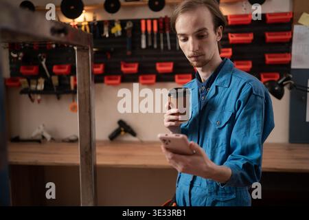 Young craftsman in overalls drinking coffee and using smartphone during break. copy space Stock Photo