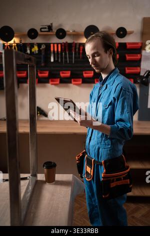 Male repairman wearing blue overalls using digital tablet for technical planning or ordering equipment in professional workshop. Repair production and Stock Photo