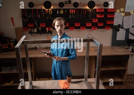 Young professional craftsman in blue overalls holding and using a digital tablet while standing in an organized workshop with tools hanging on the wal Stock Photo