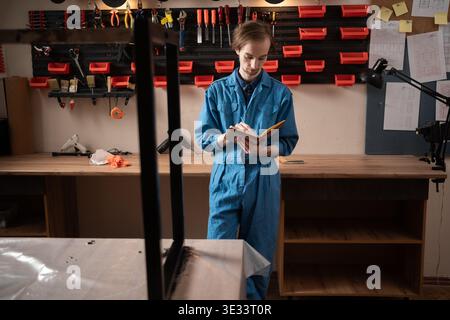 Young professional craftsman in blue overalls making notes in notebook while standing near workbench with tools hanging on wall and metal frame. Stock Photo