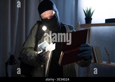 Thief wearing black balaclava holding book, flashlight and showing dollar bills standing in dark room at night. Robbery, theft and crime concept Stock Photo