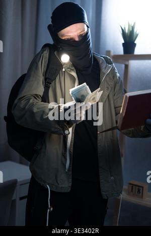 Thief wearing black balaclava holding book, flashlight and showing dollar bills standing in dark room at night. Robbery and crime concept Stock Photo