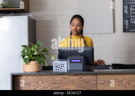 African American woman in mustard top and apron, serving behind counter in small cafe, tapping POS Stock Photo