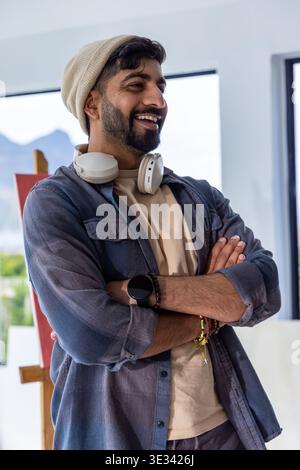 Bearded man standing, smiling and looking right in studio wearing beanie and headphones near easel Stock Photo
