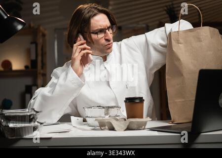 male doctor ordering takeaway food calling into restaurant using smartphone at hospital while working on night shift. Concept of consumption, medicine Stock Photo