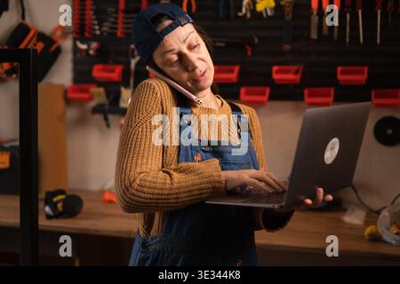 Female mechanic provides customer service via video call on a laptop in her garage workshop, managing a metal furniture repair contract. Tech, remote. Stock Photo