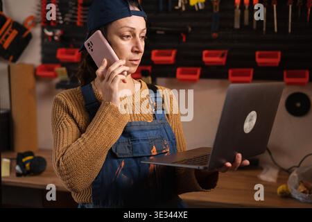 Entrepreneur in blue overalls calls a client on her laptop, discussing furniture restoration details in her small business metalworking studio. Sales, Stock Photo