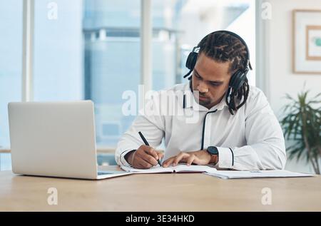 Businessman, writing and headphones with laptop or book in office for schedule, music or audio. Man, employee or taking notes with notebook, computer Stock Photo