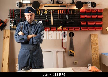 Sad tired young carpenter puffing cheeks to assemble furniture working in studio or workshop with tools on walls. woodworking workshop. looking at cam Stock Photo