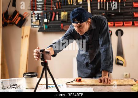 Skilled carpenter in wood workshop recording video for social media and streaming live working process. Male joiner filming DIY tutorial or worker blo Stock Photo