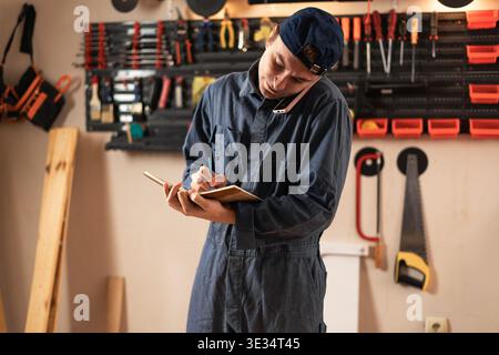 Handyman taking notes in notebook while talking with client on mobile phone in carpentry workshop. Male professional worker or carpenter discussing or Stock Photo