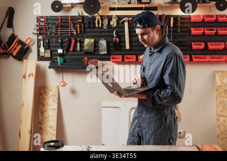 Small business owner craftsman in workwear using laptop computer in his workshop for online shopping. Male artisan or carpenter ordering tools or mate Stock Photo