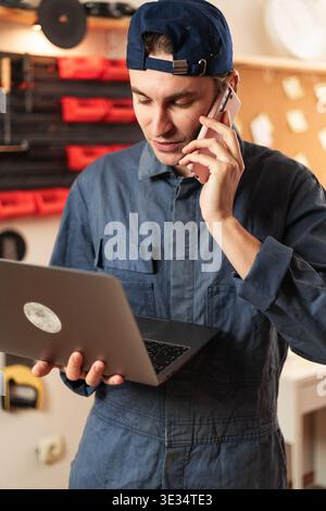 Small business owner carpenter talking on phone while using laptop in woodworking workshop. Male artisan discussing order with client and buying mater Stock Photo